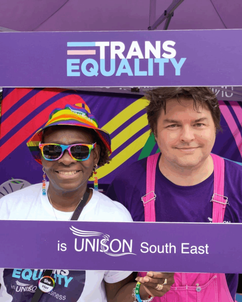 Two UNISON activists smiling for the camera with a placard which says 'Trans Equality is UNISON South East'