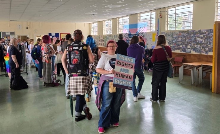 A person stands in a busy hall holding a sign that reads 'Trans Rights are Human Rights'