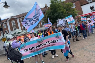 A group of people marching outside holding various pro-Trans signs, including a banner which reads 'Trans Pride Surrey'